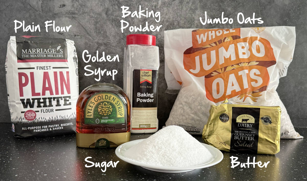 Bags and containers of plain flour, golden syrup, baking powder, jumbo oats, butter and sugar on a plate.