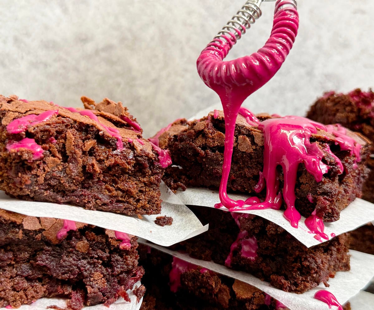 A whisk drizzling pink icing over a stack of beetroot brownies.